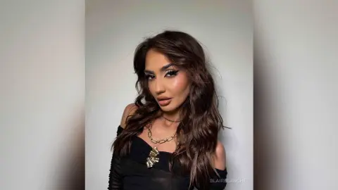 BlairBlanche poses with long wavy brown hair and winged eyeliner, wearing a black off-the-shoulder top and a gold chain necklace.