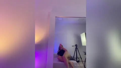 AlicehCruz poses naked on a bed, wearing a black bodysuit while looking into a mirror in a room with soft lighting.