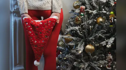 Woman in red leggings holding a festive red Santa hat next to a decorated Christmas tree in a cozy home setting.