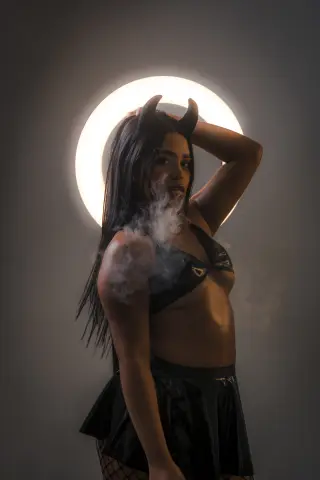 Mysterious woman in devil horns and black bikini top posing with smoke against a glowing circular light.