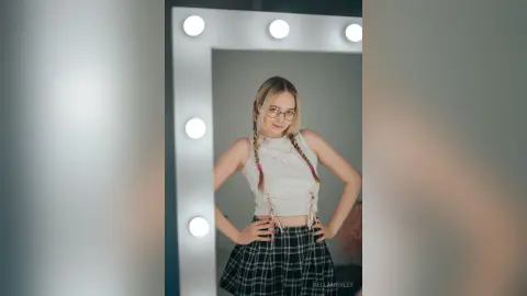 Young woman with braided hair wearing a white crop top and plaid skirt posing in a lighted vanity mirror.