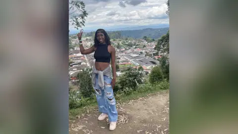 NatyGlow smiles and flashes a peace sign, wearing a black crop top and ripped blue jeans while standing outdoors overlooking a town.