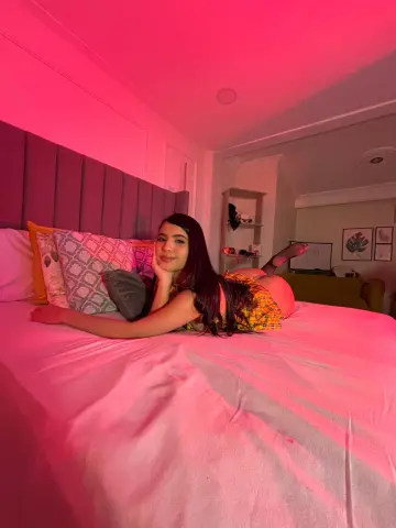Smiling woman in a yellow plaid outfit relaxing on a bed in a cozy room with pink ambient lighting.