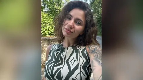 Smiling woman in a green and white patterned sleeveless top posing outdoors in a lush garden setting.