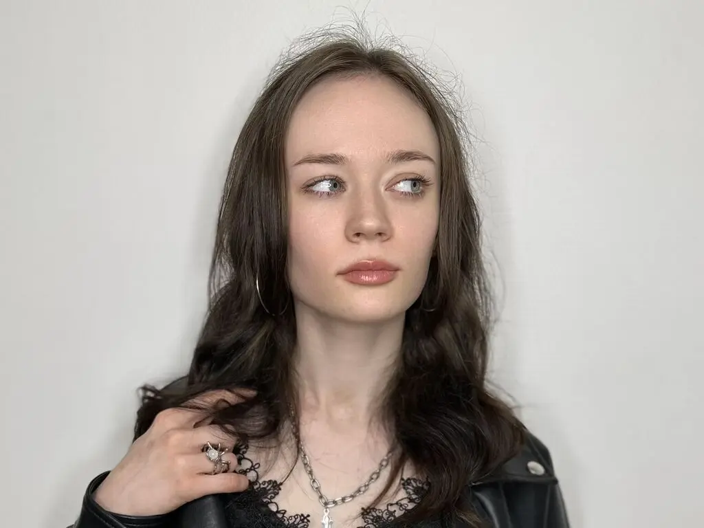 Young woman with long wavy hair wearing a black lace top and silver necklace in a minimalist studio setting.