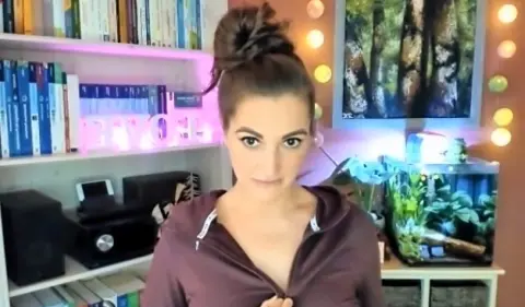 AntoniaPur28 looks at the camera with her dark hair in a high bun, wearing a dark maroon zip-up hoodie in her bedroom setting.