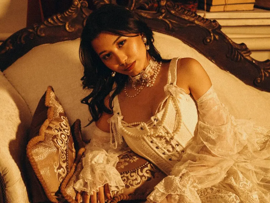 Elegant woman in a white lace corset and pearl necklace relaxing on a vintage sofa in warm, moody lighting.