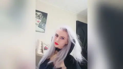 Woman with long silver hair in a black gothic outfit posing in a bright room.