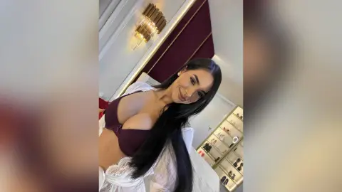 Alison smiles while wearing a maroon bra and white lace lingerie, showing her long black hair and cleavage.
