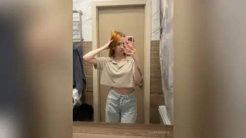 Young woman in a beige crop top and light denim jeans taking a mirror selfie in a modern bathroom.