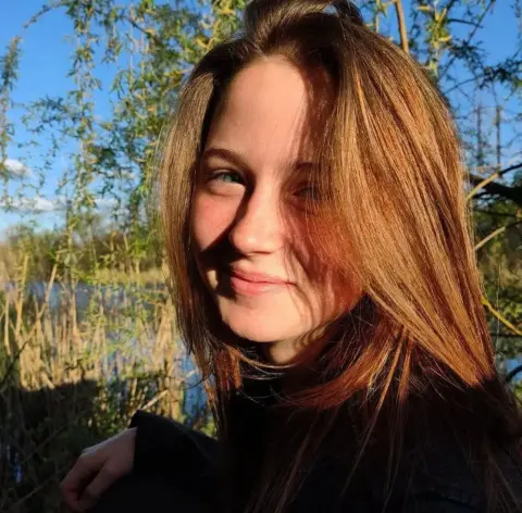 Smiling young woman with long brown hair in a black outfit outdoors in a sunny natural park setting.