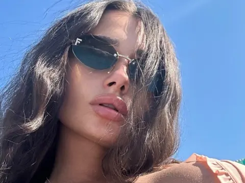 Close-up of a woman with long dark hair wearing stylish sunglasses under a bright blue sunny sky.