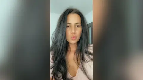 Young woman with long dark hair wearing a pink top making a playful kissy face in a bright indoor setting.