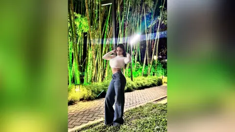 MiaKorz poses outdoors at night in a white long-sleeve crop top and dark denim flare jeans, standing in front of a lush bamboo forest.