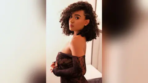 Elegant woman with curly hair wearing a black lace off-the-shoulder dress in a soft, minimalist indoor setting.