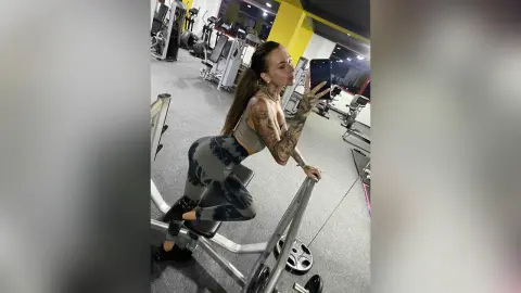 Tattooed woman in grey gym wear sitting on a weight machine in a modern fitness center.
