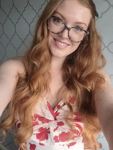 Anna_OG smiles wearing glasses and a red and white floral print top, showing off her long, wavy red hair against a patterned wall.