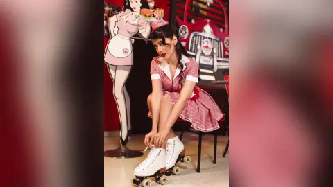 JessieLiu wears a red and white checkered waitress outfit and white roller skates while sitting in a diner setting.