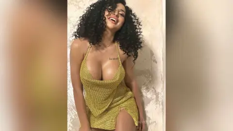 NicoleBella smiles while wearing a low-cut yellow mesh dress that shows off her cleavage and chest.