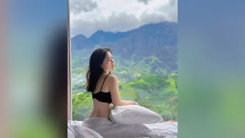Young woman in black lingerie relaxing in bed overlooking a lush, misty mountain landscape at sunrise.