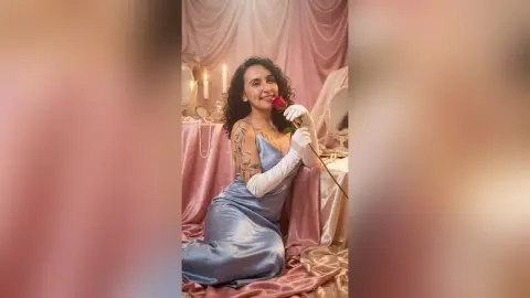 Smiling woman in a blue satin dress and white gloves holding a red rose in a romantic, vintage setting.