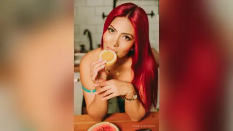IsabellaEstrada with long red hair licks a round slice of citrus fruit while leaning forward on a wooden table.
