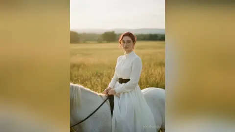 Redheaded woman in a white long-sleeve dress riding a white horse through a golden sunlit meadow.