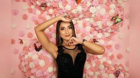 Smiling woman in a black lace dress posing against a pink rose and butterfly floral backdrop.