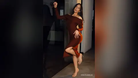 MarcelaMilan poses in a tight, long-sleeved dark red dress and high heels, leaning against a glass door.