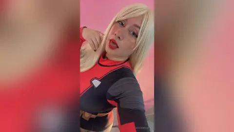 Blonde woman in a red and black superhero cosplay outfit posing against a pink background.