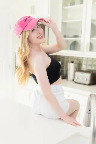Smiling blonde woman in a pink cap and black crop top sitting on a white kitchen counter in a bright home.