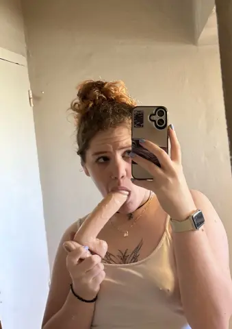 Young woman in a white tank top taking a playful mirror selfie in a bright room.