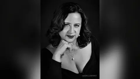 IsabellaBennet poses with her hand under her chin, wearing a low-cut black top and a pearl necklace against a dark background.