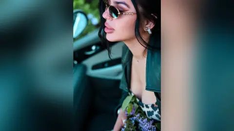 Stylish woman in sunglasses and a black leather jacket holding flowers inside a car on a sunny day.