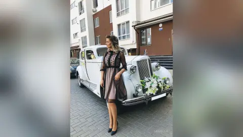 Elegant woman in a black sheer dress standing by a vintage white car on a city street.