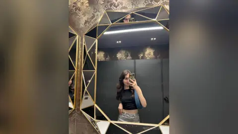 Young woman in a black crop top and silver pants taking a mirror selfie in a luxury gold-trimmed room.