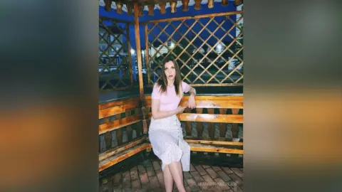 Woman in a pink top and white floral skirt sitting in a wooden gazebo at twilight.