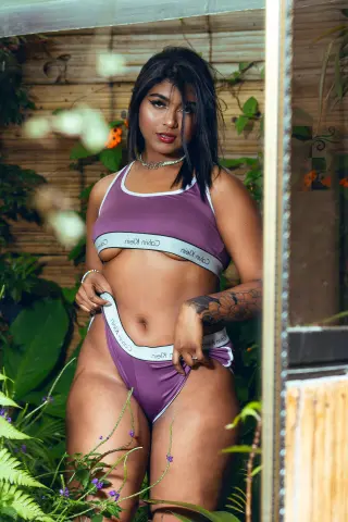 Young woman in purple Calvin Klein lingerie posing outdoors in a lush green garden setting.