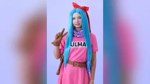 Cosplayer in pink Ulma outfit with long blue hair and red bow posing playfully against a light background.