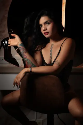 Tattooed woman in a black lace bodysuit sitting in a moody, dimly lit room holding a black high heel shoe.