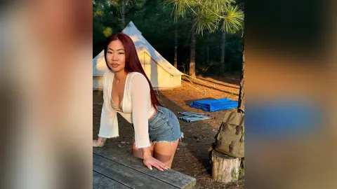 Red-haired woman in a white blouse and denim shorts relaxing by a white tent at a sunny forest campsite.