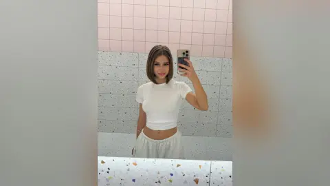 Young woman in a white t-shirt and grey lounge pants taking a mirror selfie in a pink tiled bathroom.