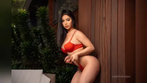VickyPortman poses against a wooden wall wearing a red bra and matching red thong or bikini bottom.
