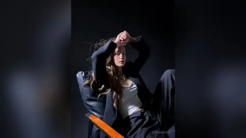 LissaDesso poses in a black blazer and white tank top with her hands behind her head against a dark background.