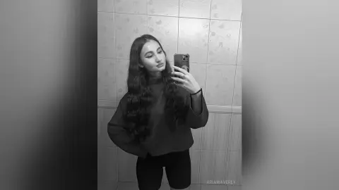 Young woman in a dark long-sleeve top taking a moody black and white mirror selfie in a tiled bathroom.