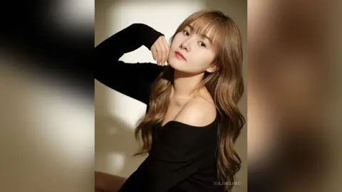XiaJiaojiao poses with her hands near her face, wearing a black off-the-shoulder top with long wavy hair against a plain background.