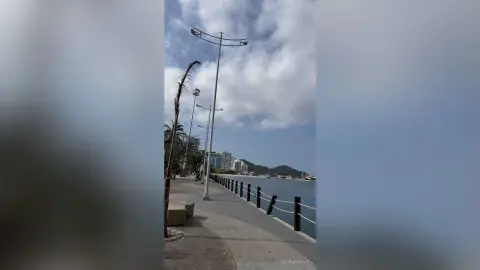Empty concrete boardwalk with street lamps overlooking a calm ocean under a cloudy blue sky.