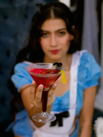 SusanaSanchez20 holds a red cocktail in a martini glass, wearing a blue and white maid outfit with dark hair and red lipstick.