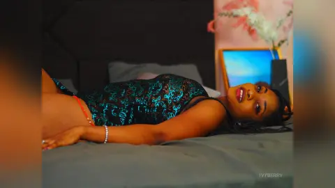IvyBerry lies on a bed wearing a black and teal lace bodysuit, looking toward the camera with her dark hair spread out.