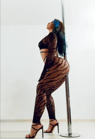 Curvy woman in a black sheer patterned bodysuit posing by a silver pole in a bright minimalist studio.